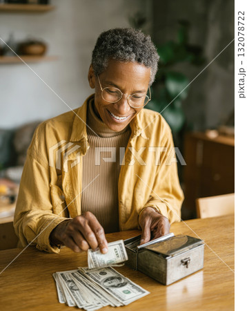 A warm, vertical portrait of a smiling mature woman sitting at wooden table, counting and putting bills into a vintage metal box or safe. Image conveys themes of saving, retirement, financial security 132078722