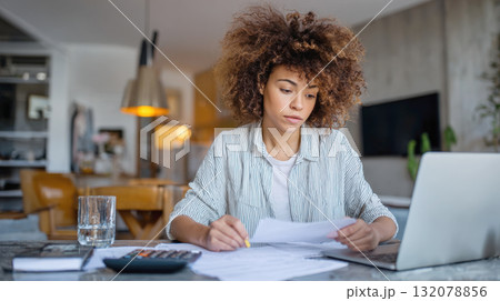 woman with striking curly hair reviews bills, financial documents at home, using laptop, calculator. Scene conveys concepts of personal finance management, tax preparation, remote work, paying bills. 132078856