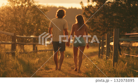 Romantic rear view of a young couple walking barefoot down a grassy path, holding hands during sunset. Scene evokes summer love, freedom, nature connection, serene country living, vacation getaway Romantic rear view of a young couple walking barefoot down a grassy path, holding hands during sunset. Scene evokes summer love, freedom, nature connection, serene country living, vacation getaway 132078895