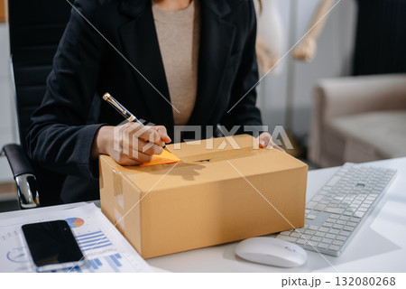 Asian woman prepares parcels and checks online orders on tablet and laptop for delivery online shopping and small business success Asian woman prepares parcels and checks online orders on tablet and laptop for delivery online shopping and small business success 132080268
