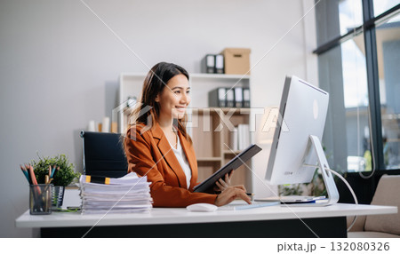 Young Asian woman, smiling and working at her desk in an office environment. She is wearing a business suit using a laptop. Young Asian woman, smiling and working at her desk in an office environment. She is wearing a business suit using a laptop. 132080326