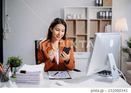 Young Asian woman, smiling and working at her desk in an office environment. She is wearing a business suit using a laptop. 132080341