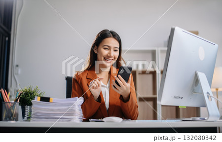 Young Asian woman, smiling and working at her desk in an office environment. She is wearing a business suit using a laptop. Young Asian woman, smiling and working at her desk in an office environment. She is wearing a business suit using a laptop. 132080342