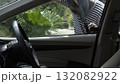 masked individual wearing a striped shirt using a metal tool to unlock the car through the window seal in daylight with greenery in the background 132082922