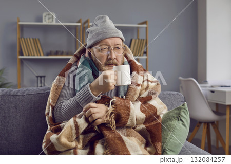 Senior man sitting wrapped in a blanket and trying to warm himself with a cup of hot tea. 132084257