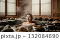 Asian woman relaxing in traditional stone hot spring bath with steaming water surrounded by ceramic bowls containing natural herbs and spices in korean spa setting 132084690