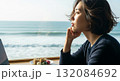 Asian woman with short bob haircut gazing thoughtfully out window at ocean waves while working on laptop with traditional desserts on wooden table 132084692