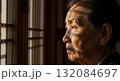 Elderly Asian woman in profile gazing pensively through traditional wooden window frame with warm golden sunlight illuminating weathered facial features 132084697