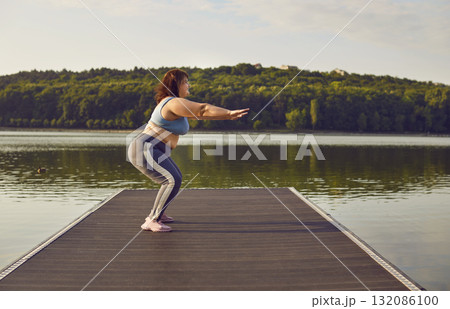 Overweight fat woman in sportswear doing sport exercises standing on the pier in summer park. 132086100