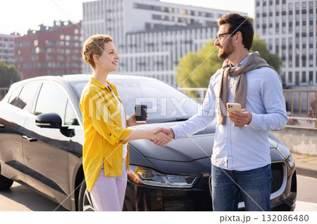 A man and a woman shake hands near a sleek black electric car, possibly after a business deal or meeting. 132086480