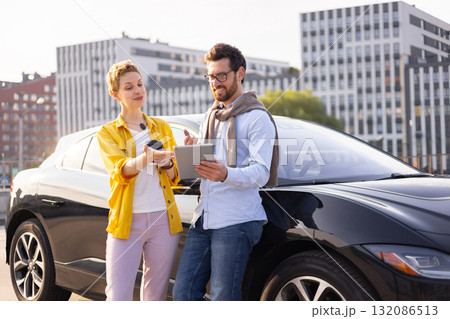 A man and woman are looking at a tablet next to a black car, possibly discussing business or planning a trip. A man and woman are looking at a tablet next to a black car, possibly discussing business or planning a trip. 132086513