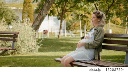 Happy pregnant woman sitting outside touching her belly calming the baby. Happy pregnant woman sitting outside touching her belly calming the baby. 132087294