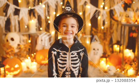 A happy young boy dressed in a skeleton costume and witch hat smiles brightly, celebrating Halloween. He's surrounded by warm, festive decorations like string lights, pumpkins, and candles A happy young boy dressed in a skeleton costume and witch hat smiles brightly, celebrating Halloween. He's surrounded by warm, festive decorations like string lights, pumpkins, and candles 132088354