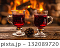 Two elegant glass mugs filled with steaming red beverage sit on a rustic wooden table, with a cozy fireplace glowing warmly in the background, creating a festive atmosphere 132089720