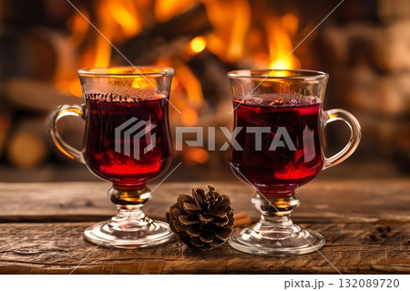 Two elegant glass mugs filled with steaming red beverage sit on a rustic wooden table, with a cozy fireplace glowing warmly in the background, creating a festive atmosphere 132089720