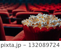 Close-up of a red popcorn bucket filled with buttery popcorn, set against a blurred background of empty red theater seats, evoking a cinematic experience and anticipation 132089724