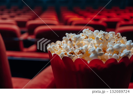 Close-up of a red popcorn bucket filled with buttery popcorn, set against a blurred background of empty red theater seats, evoking a cinematic experience and anticipation 132089724