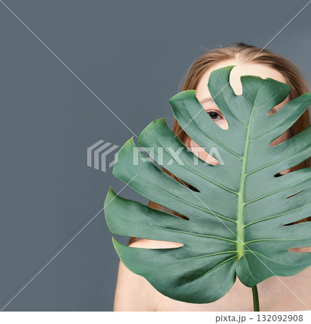 Caucasian female young adult partially concealed by large monstera leaf against 132092908