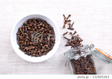 Dried cloves, in a white bowl, on linen. Aromatic flower buds of the tree Syzygium aromaticum, used as spice, fragrance, and for flavoring in cuisine and cosmetic products. Close-up, from above. Photo 132093263
