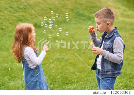 Young caucasian children blowing bubbles outdoors on grass in denim outfits Young caucasian children blowing bubbles outdoors on grass in denim outfits 132093368