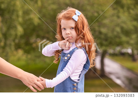 Red-haired caucasian young girl with blue bow in denim dress holding adult hand 132093383