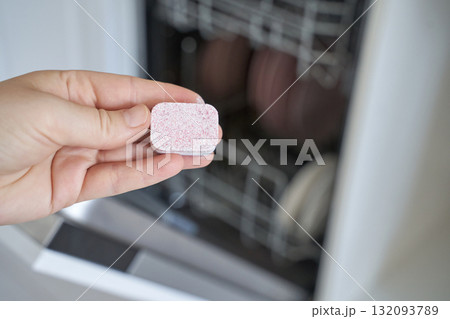 Hand holding dishwasher tablet in kitchen with open dishwasher in background 132093789