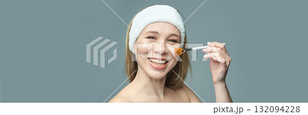 Smiling caucasian female using makeup brush in skincare routine on blue Smiling caucasian female using makeup brush in skincare routine on blue 132094228