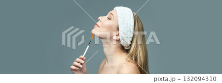 Young caucasian female with headband applying makeup with brush on chin, side 132094310