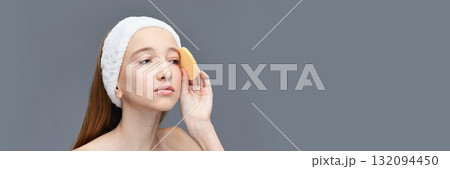 Young caucasian female cleansing face with sponge and headband on gray Young caucasian female cleansing face with sponge and headband on gray 132094450