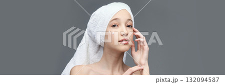 Young caucasian female applying skincare cream with towel on head against gray 132094587