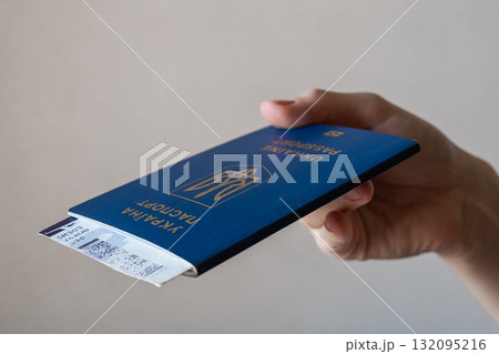 A hand holding a blue Ukrainian passport. The passport features the national emblem and text in Ukrainian and English. The background is neutral and simple. 132095216