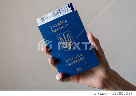 A hand holding a blue Ukrainian passport. The passport features the national emblem and text in Ukrainian and English. The background is neutral and simple. 132095217