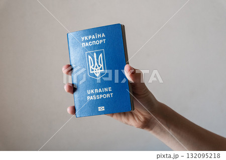 A hand holding a blue Ukrainian passport. The passport features the national emblem and text in Ukrainian and English. The background is neutral and simple. 132095218
