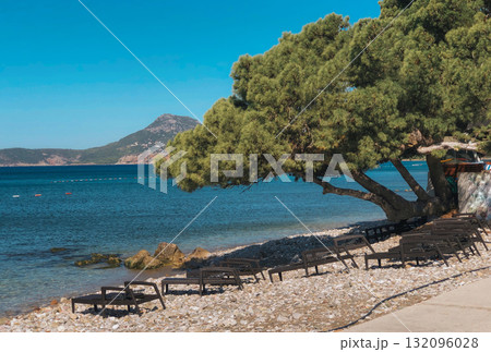 A bright, sunny, wide-angle photograph capturing a tranquil scene on a Mediterranean beach in a coastal resort area, likely in Montenegro or Croatia. 132096028