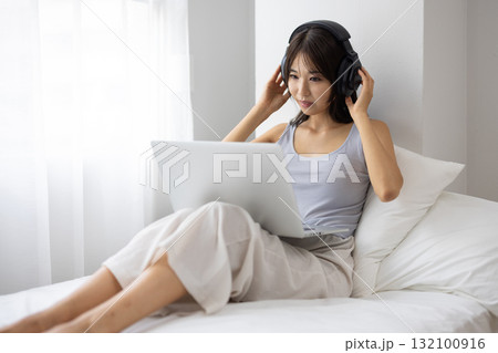 ベッドの上でヘッドフォンをつけてパソコンを操作する若い女性 ベッドの上でヘッドフォンをつけてパソコンを操作する若い女性 132100916