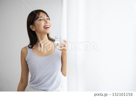 窓辺で水が入ったグラスを持ち微笑む若い女性 窓辺で水が入ったグラスを持ち微笑む若い女性 132101158