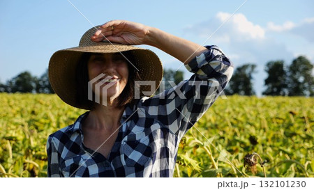 Happy smiling female farmer looks into camera standing near sunflower field. Portrait of adult beautiful agronomist in straw hat with yellow plantation at background. Agricultural business concept Happy smiling female farmer looks into camera standing near sunflower field. Portrait of adult beautiful agronomist in straw hat with yellow plantation at background. Agricultural business concept 132101230