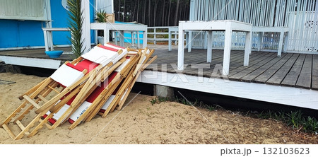 Neatly stacked red and white wooden lounge chairs on a sandy terrace next to a beach hut. Concept of the end of the beach season, a small business in operation Neatly stacked red and white wooden lounge chairs on a sandy terrace next to a beach hut. Concept of the end of the beach season, a small business in operation 132103623