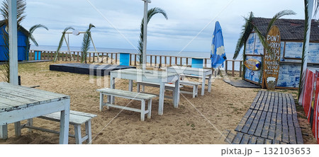 Closed at the end of the season, this coastal beach bar features white tables, palm tree decor, and a surfboard-shaped sign by the ocean. 132103653