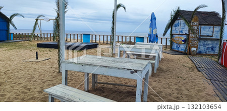Closed at the end of the season, this coastal beach bar features white tables, palm tree decor, and a surfboard-shaped sign by the ocean. 132103654