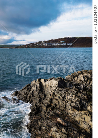 Rough waves crash against the rocky coast of Inchydoney Island. Houses sit on the hillside in the distance under an overcast sky. The ocean stretches out to the horizon. 132104407