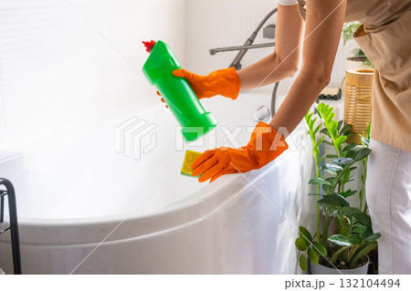 Woman in rubber gloves and apron manually washes bathtub is in bathroom of house with sponge cleaner and cleaning agent in a bottle. Restoring order, cleanliness and hygiene in home, cleaning servise Woman in rubber gloves and apron manually washes bathtub is in bathroom of house with sponge cleaner and cleaning agent in a bottle. Restoring order, cleanliness and hygiene in home, cleaning servise 132104494