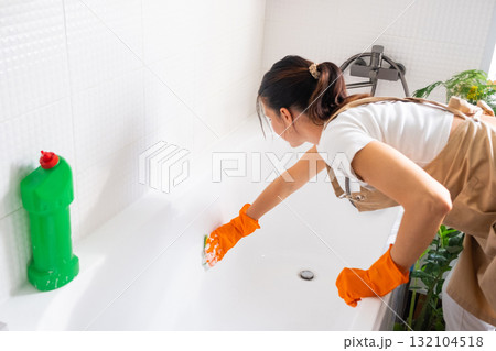 Woman in rubber gloves and apron manually washes bathtub is in bathroom of house with sponge cleaner and cleaning agent in a bottle. Restoring order, cleanliness and hygiene in home, cleaning servise 132104518