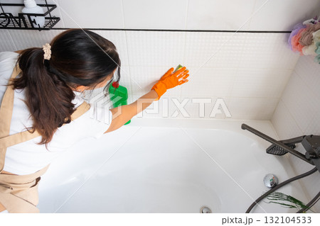 Woman in rubber gloves and apron manually washes bathtub is in bathroom of house with sponge cleaner and cleaning agent in a bottle. Restoring order, cleanliness and hygiene in home, cleaning servise 132104533