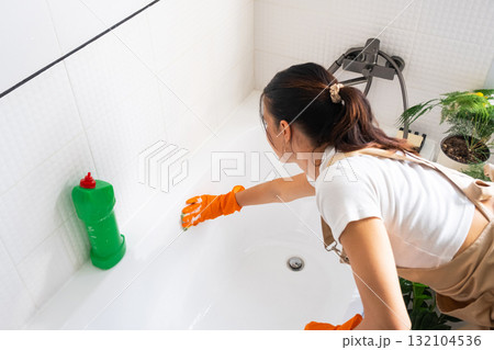 Woman in rubber gloves and apron manually washes bathtub is in bathroom of house with sponge cleaner and cleaning agent in a bottle. Restoring order, cleanliness and hygiene in home, cleaning servise 132104536