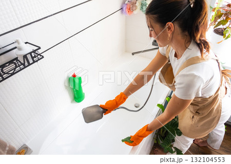 Woman in rubber gloves and apron manually washes bathtub is in bathroom of house with sponge cleaner and cleaning agent in a bottle. Restoring order, cleanliness and hygiene in home, cleaning servise Woman in rubber gloves and apron manually washes bathtub is in bathroom of house with sponge cleaner and cleaning agent in a bottle. Restoring order, cleanliness and hygiene in home, cleaning servise 132104553