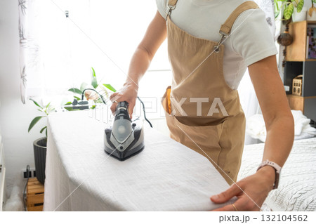 A housewife woman in an apron irons linen curtains in a room on an ironing board A housewife woman in an apron irons linen curtains in a room on an ironing board 132104562