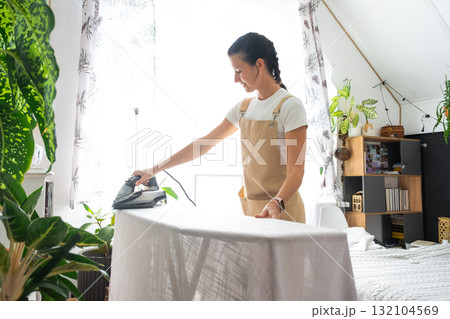 A housewife woman in an apron irons linen curtains in a room on an ironing board A housewife woman in an apron irons linen curtains in a room on an ironing board 132104569