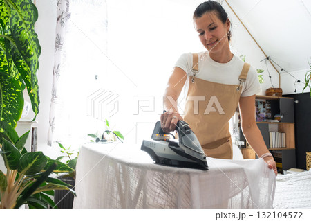 A housewife woman in an apron irons linen curtains in a room on an ironing board A housewife woman in an apron irons linen curtains in a room on an ironing board 132104572