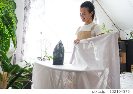 A housewife woman in an apron irons linen curtains in a room on an ironing board A housewife woman in an apron irons linen curtains in a room on an ironing board 132104576